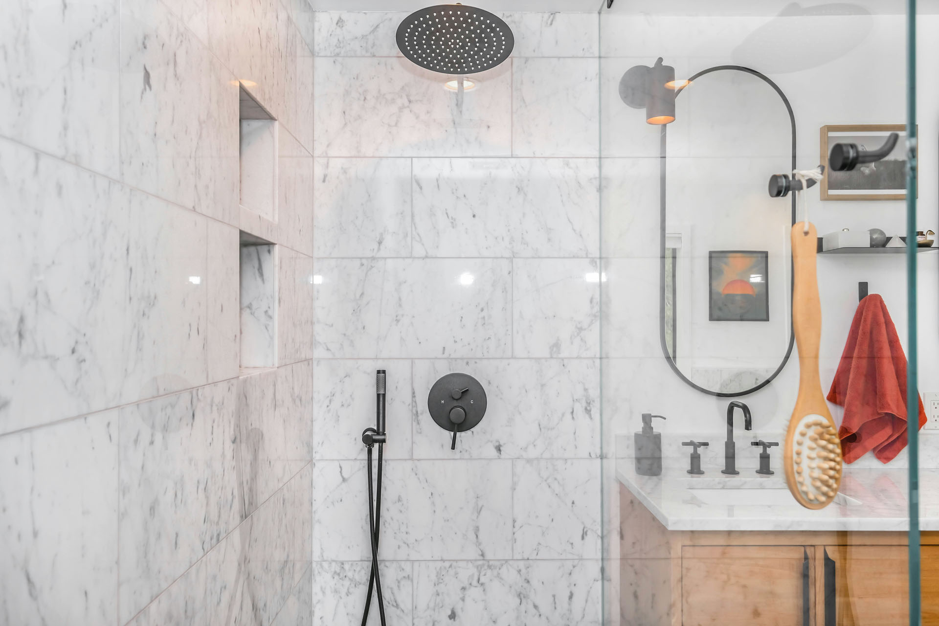 Modern bath with marble shower walls, black fixtures, a rain showerhead, vanity with rectangular mirror, and a hanging wooden bath brush. A red towel hangs on the wall, creating a stylish wet space by Trubath.