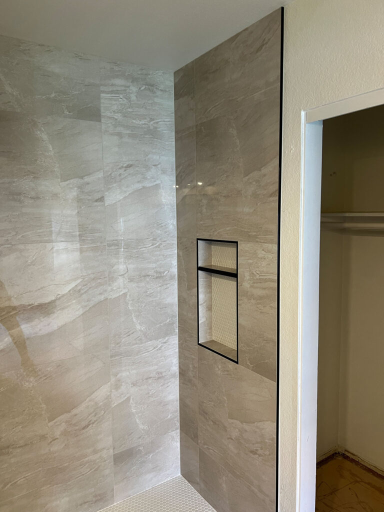 Shower with large beige tiles, a recessed shelf with black trim, and a view of an adjacent closet with a hanging rod—perfect inspiration for your next bathroom remodel Phoenix project.