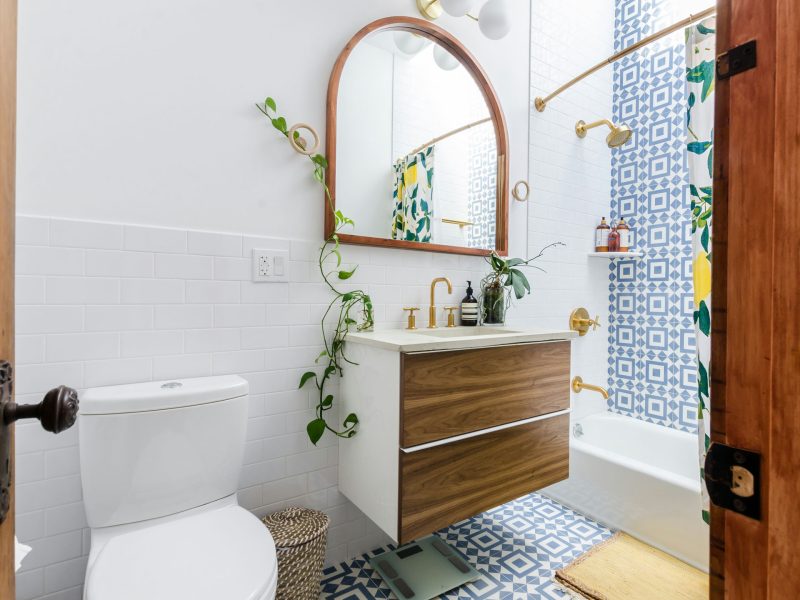 Modern bathroom remodel with a white toilet, floating wood vanity, large round mirror, potted plants, and blue patterned Trubath tiles in the shower area.