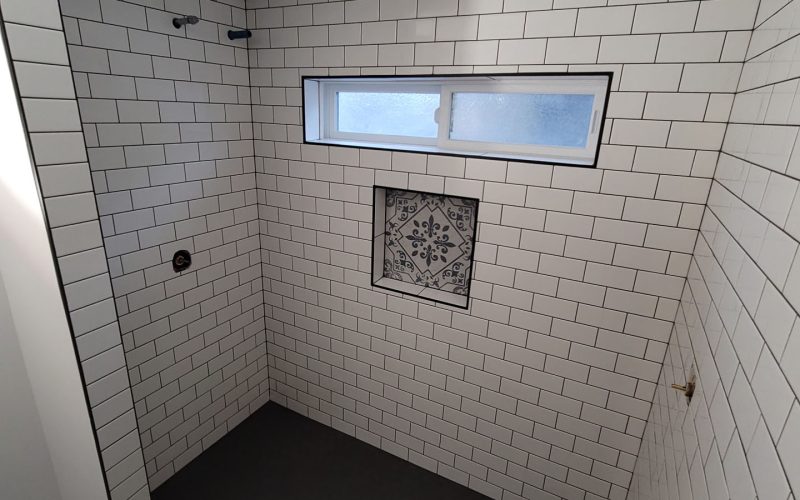 A walk-in shower by Trubath features white subway tile walls, a decorative tile niche, a long horizontal window, and dark hexagonal floor tiles—perfect for your wet space remodel.